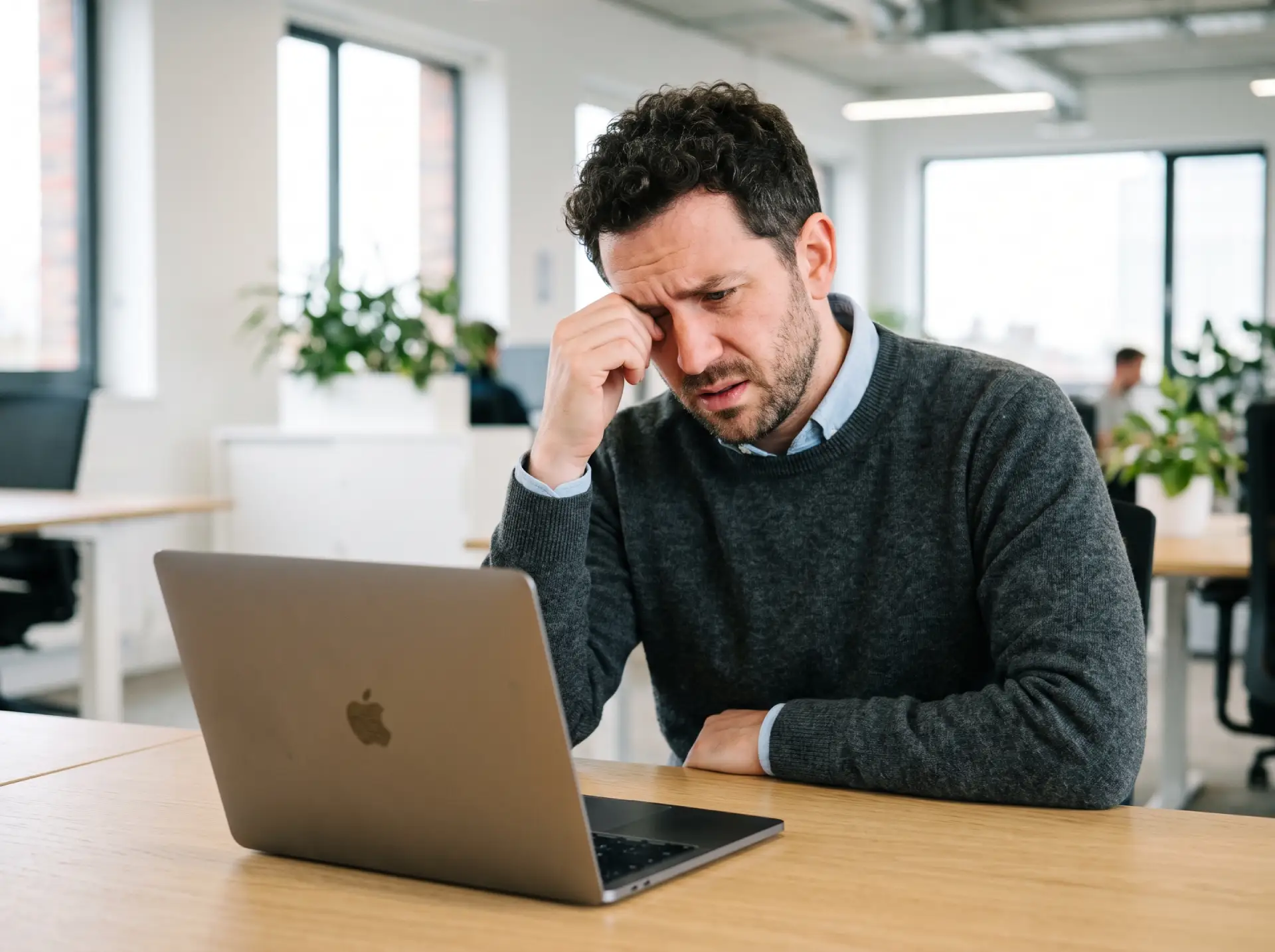 A stressed man rubbing his eye in frustration while looking at a laptop screen in a bright, modern office.